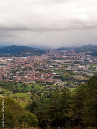 A city view with a cloudy sky and mountains in the background. The city is full of buildings and trees