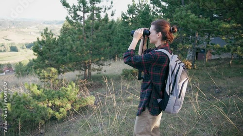 A young Caucasian woman uses binoculars to observe birds while walking through the autumn forest