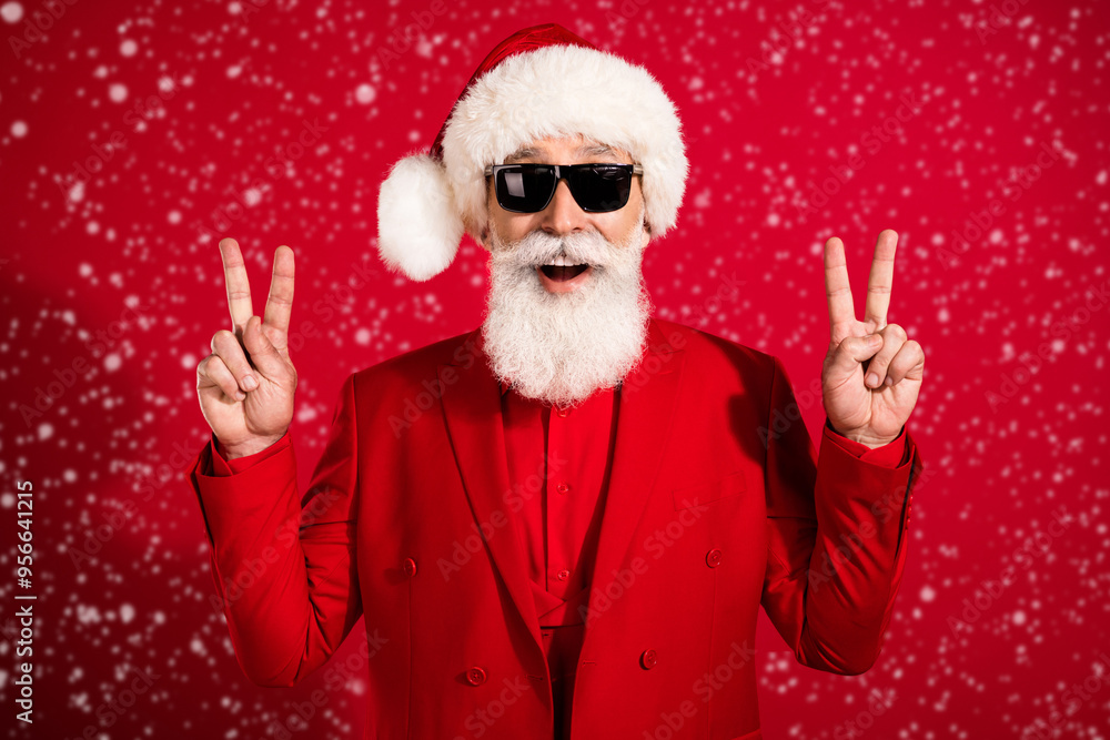 Photo of old positive happy man make fingers v-signs wear santa claus hat isolated on red color background Photo of old positive happy man make fingers v-signs wear santa claus hat isolated on red color background