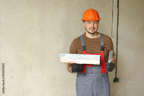 repairman in overalls and protective orange helmet holds two spatulas for plastering concrete wall with putty using. Repair work at apartment and construction new building concept. Copy space