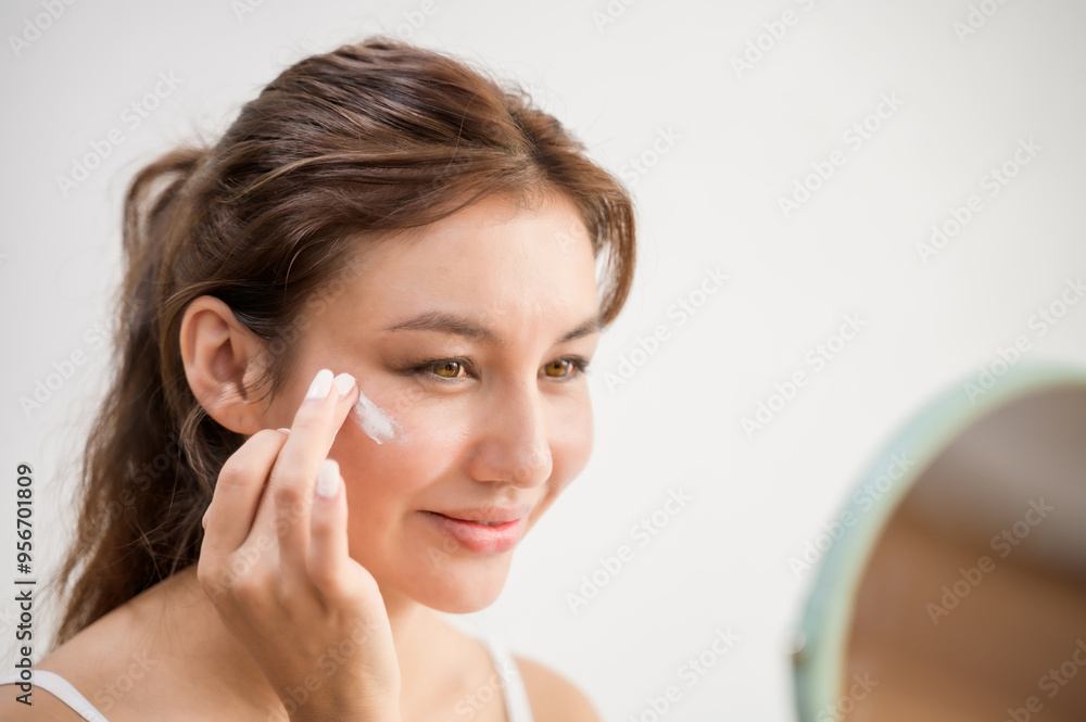 Close up portrait photo of young beautiful Asian woman taking care of her facial skin apply face skin care cosmetic lotion moisturizer product and checking in mirror at home. Beauty care concept Close up portrait photo of young beautiful Asian woman taking care of her facial skin apply face skin care cosmetic lotion moisturizer product and checking in mirror at home. Beauty care concept