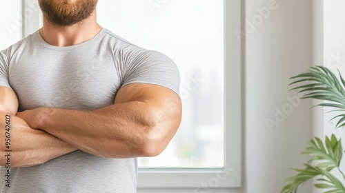 A confident man with muscular arms stands in a bright room, showcasing strength and wellness through a focused posture.