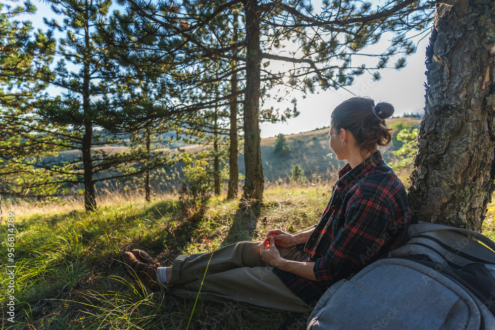Obraz premium Young female hiker sitting down to rest on the grass during a summer hiking in pine woodland