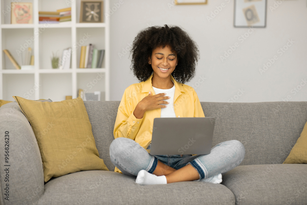 Grateful black lady putting palms on chest, being thankful to teacher during remote lesson, using laptop on sofa at home Grateful black lady putting palms on chest, being thankful to teacher during remote lesson, using laptop on sofa at home