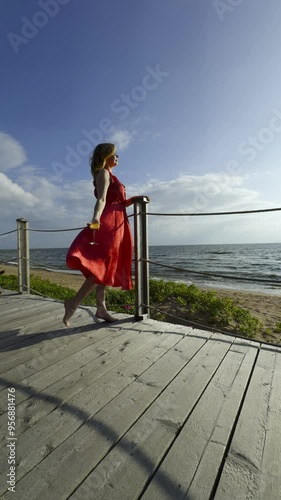 A woman in a flowing red dress dances barefoot on a wooden deck overlooking the ocean, holding a glass of champagne. The gentle breeze and the serene seaside setting capture a moment of joy and free photo