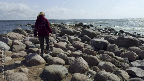 A woman wearing a red hoodie carefully navigates a rocky shoreline by the sea, with waves gently crashing against the rocks. The bright, sunny sky and the expansive ocean add a sense of adventure  photo