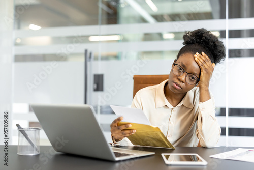 African American businesswoman feeling stressed after receiving bad news via mail at office desk. She sits with laptop and phone, reflecting on difficult situation.