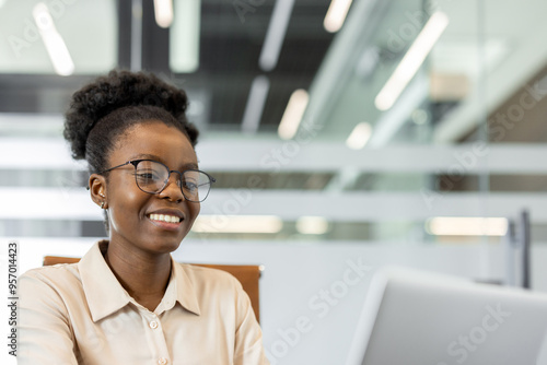 Confident African American businesswoman smiling while working on laptop in modern office setting. Represents professional success, career growth, and positive work environment.