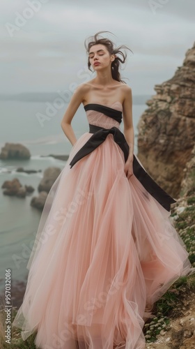 A woman in a pink dress with a black bow is standing on a rocky cliff overlookin photo