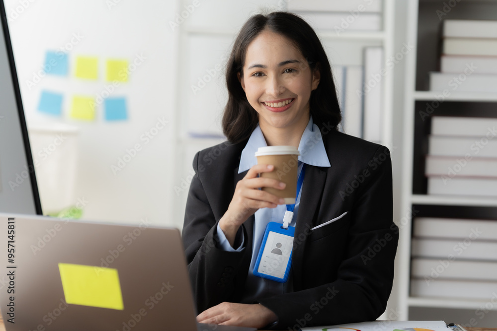 Confident Businesswoman: A smiling, confident Asian businesswoman in a professional suit sits at her desk, taking a break with a cup of coffee while working on her laptop. She exudes a sense of comp Confident Businesswoman: A smiling, confident Asian businesswoman in a professional suit sits at her desk, taking a break with a cup of coffee while working on her laptop. She exudes a sense of comp