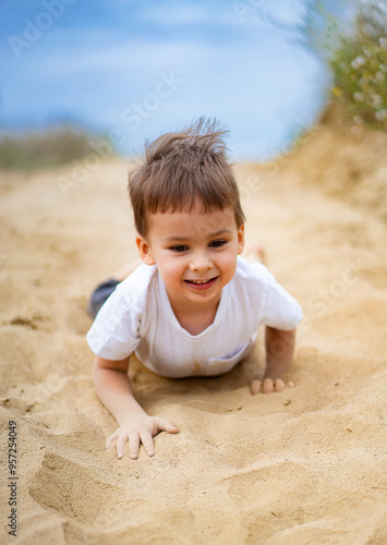 Young boy having fun crawling on sandy beach during sunny day. A cheerful young boy enjoys crawling on soft sand at the beach, smiling under the bright sun