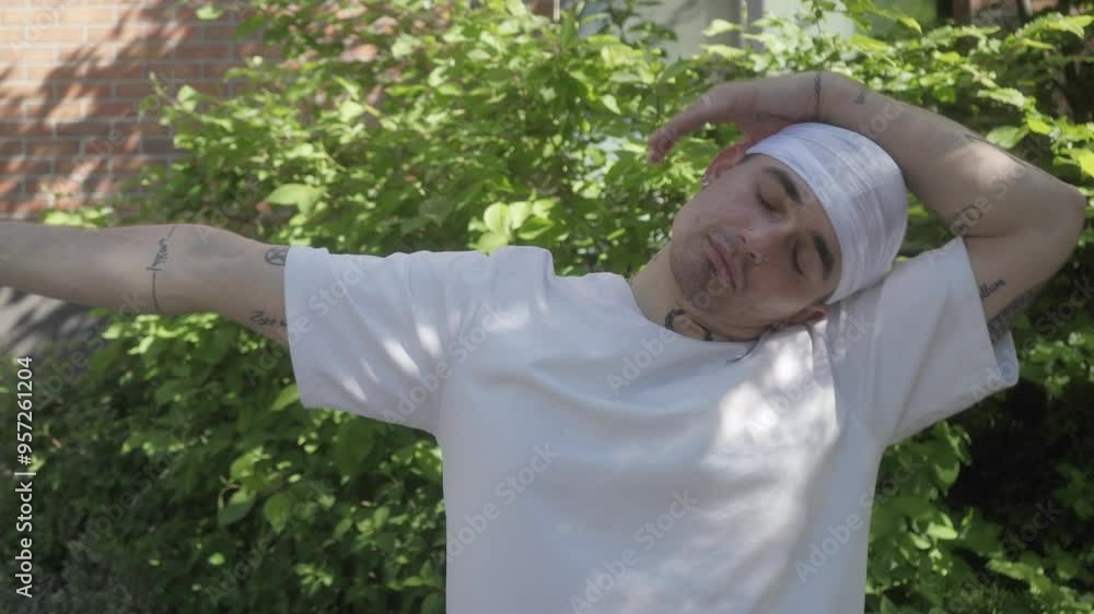 A tattooed man dancer performer wearing a white t-shirt and bandana is doing warm-up stretches in an urban setting, getting ready for a hip-hop dance routine. A tattooed man dancer performer wearing a white t-shirt and bandana is doing warm-up stretches in an urban setting, getting ready for a hip-hop dance routine.