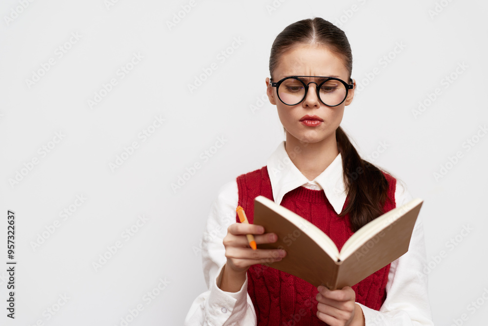 Young woman reading a book with focused expression, wearing glasses and a red vest against a plain light background Ideal for education or study themes Young woman reading a book with focused expression, wearing glasses and a red vest against a plain light background Ideal for education or study themes