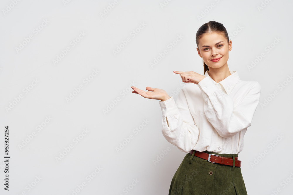 Woman pointing with a friendly smile on a neutral background, presenting something in her open palm, dressed in a white blouse and green pants Woman pointing with a friendly smile on a neutral background, presenting something in her open palm, dressed in a white blouse and green pants