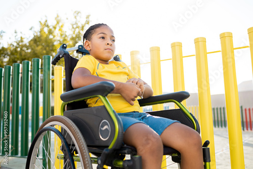 A dark-skinned boy between 8 and 9 years old is sitting in a wheelchair next to a park with a sad expression. African children with disabilities concept. photo