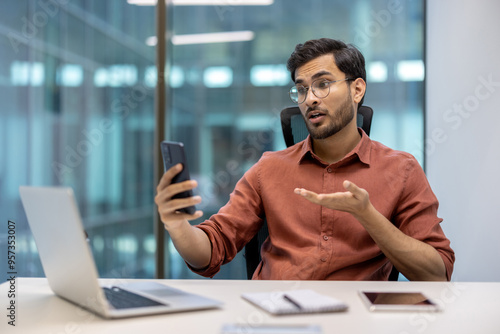 Business professional in office video conference call using smartphone and laptop, discussing work project with expressive gestures