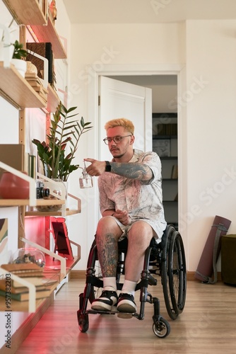 Young man with tattoos sitting in his wheelchair while watering a houseplant on shelf in modernly designed living space filled with natural light photo