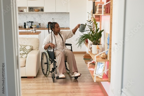 Person in wheelchair watering plants on shelf in cozy, modern apartment with wooden flooring and white furniture, creating an inclusive, homey environment photo