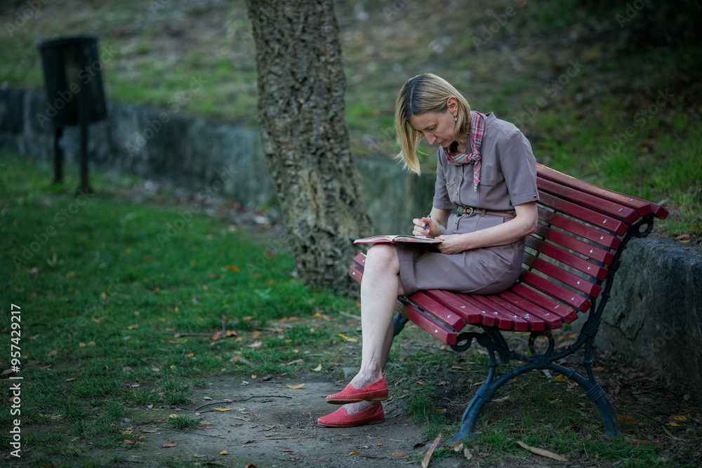 A woman sits on a park bench writing in a notebook or journal with a pen. A woman sits on a park bench writing in a notebook or journal with a pen.