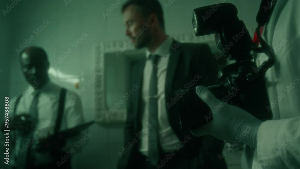 Close up view of forensic photographer in white coat and gloves holding camera and talking with two detectives standing beside in dimly lit bathroom Close up view of forensic photographer in white coat and gloves holding camera and talking with two detectives standing beside in dimly lit bathroom
