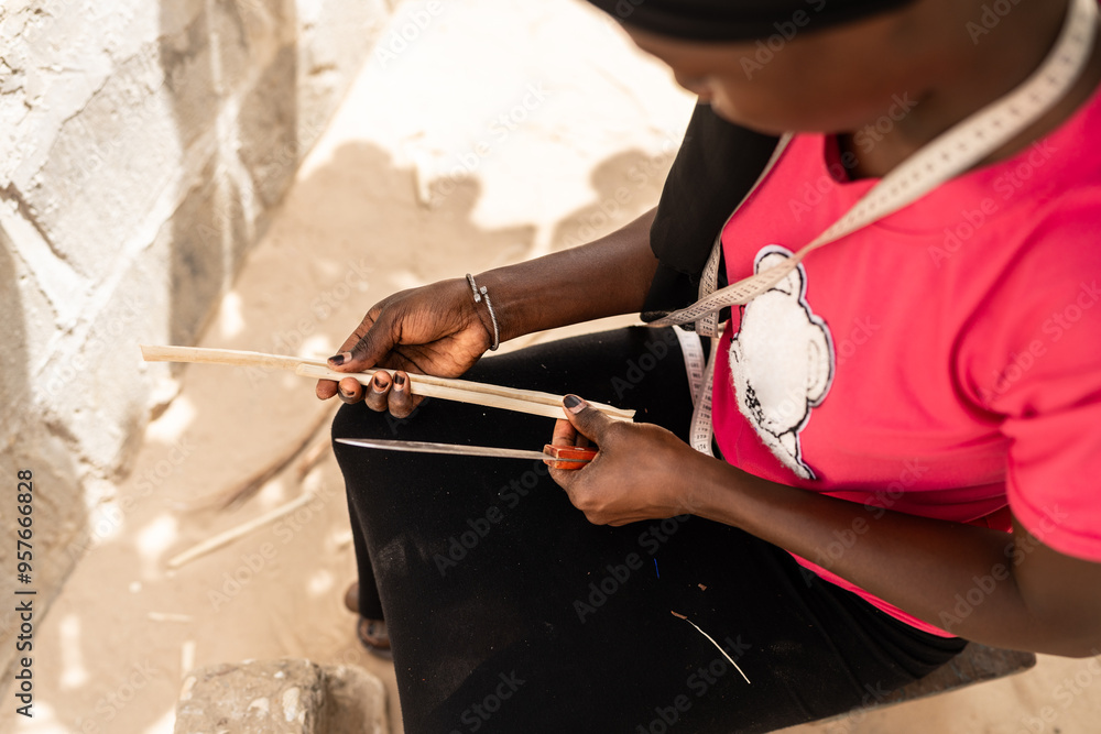 © Alvaro Lavin/Stocksy - African woman sitting and sharpening sticks for hand fan making