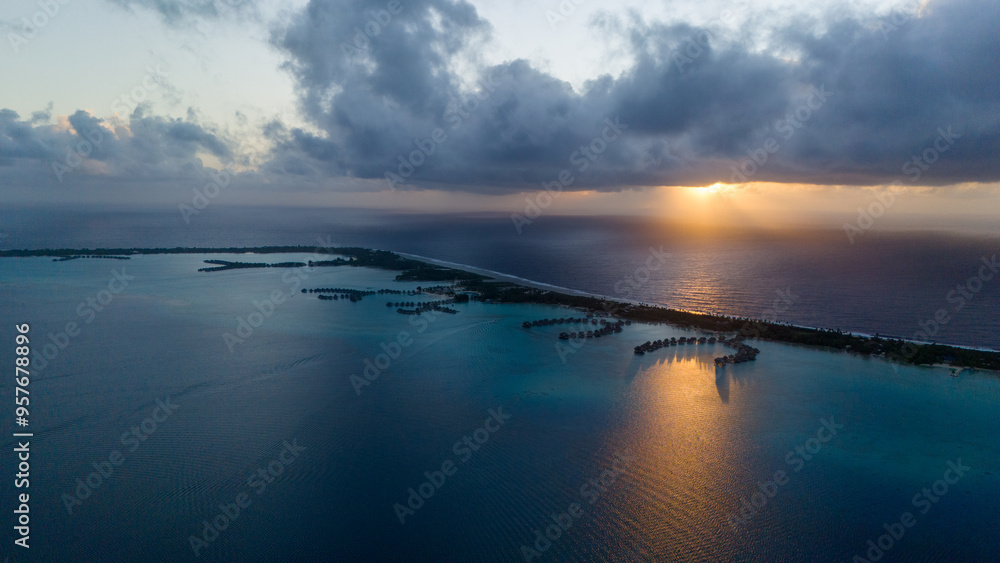 Aerial view captured by drone overlooking tropical island resorts with overwater bungalows on Bora Bora motu in French Polynesia. Azure and turquoise colored ocean sea water. Low clouds above sunrise. Aerial view captured by drone overlooking tropical island resorts with overwater bungalows on Bora Bora motu in French Polynesia. Azure and turquoise colored ocean sea water. Low clouds above sunrise.