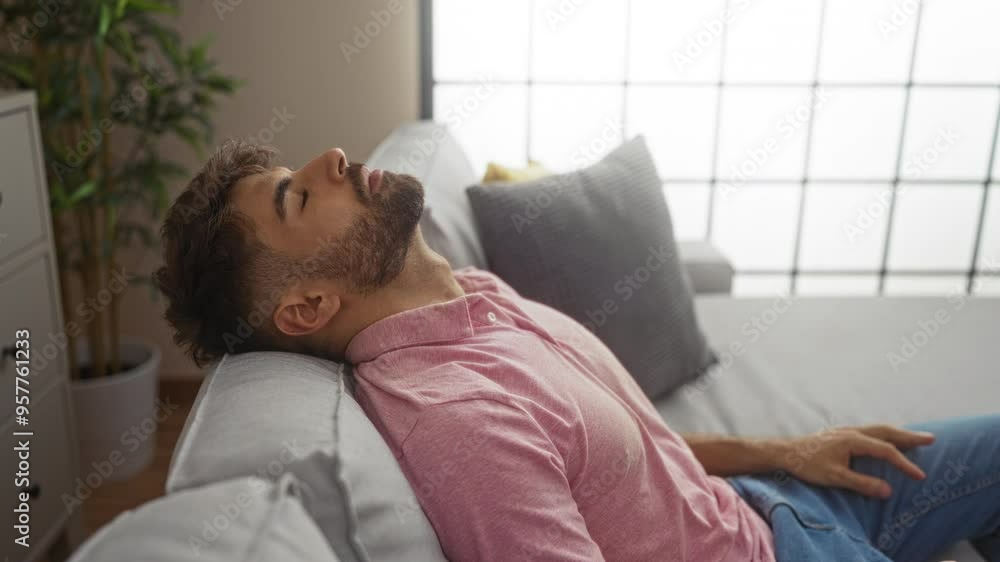 Young man relaxing in a modern living room with bright lighting and indoor plants, wearing casual pink shirt and jeans, sitting on a grey couch, captured in a peaceful, home setting Young man relaxing in a modern living room with bright lighting and indoor plants, wearing casual pink shirt and jeans, sitting on a grey couch, captured in a peaceful, home setting