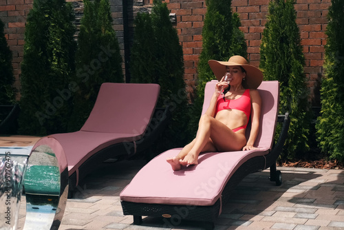 Beautiful girl in pink swimsuit and wicker hat relaxing on sun lounger near pool. Drinking water from glass. Relaxing in luxury pool