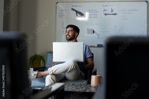 Holding laptop, Indian man smiling and working in modern robotic office photo