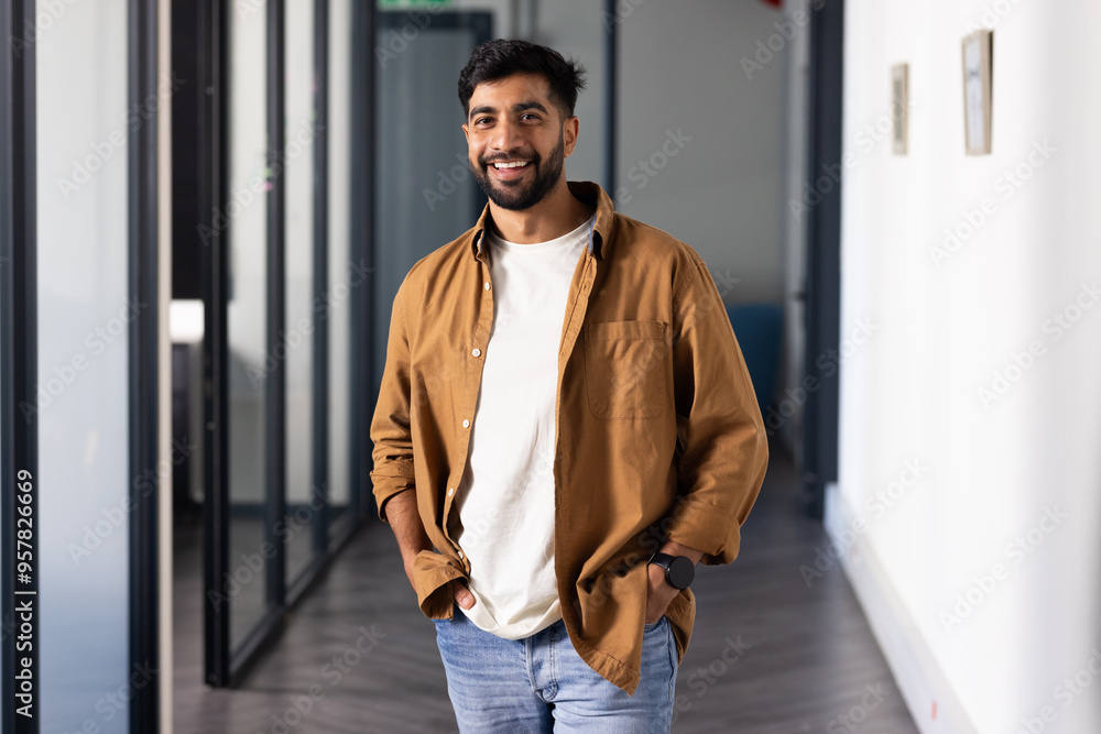Smiling Indian man in casual attire standing in modern office hallway, hands in pockets Smiling Indian man in casual attire standing in modern office hallway, hands in pockets