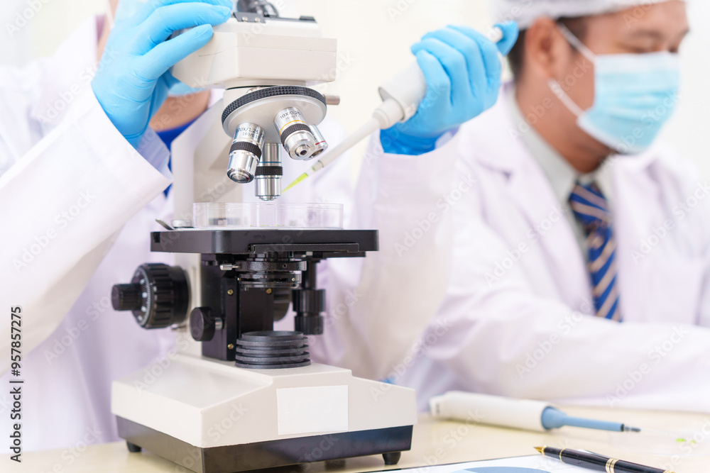 Two Asian scientists in lab coats sit at a table in a laboratory, examining samples through a microscope. The room is equipped with scientific tools, liquids, and chemistry materials. Two Asian scientists in lab coats sit at a table in a laboratory, examining samples through a microscope. The room is equipped with scientific tools, liquids, and chemistry materials.