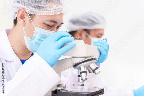 Two Asian scientists in lab coats sit at a table in a laboratory, examining samples through a microscope. The room is equipped with scientific tools, liquids, and chemistry materials.