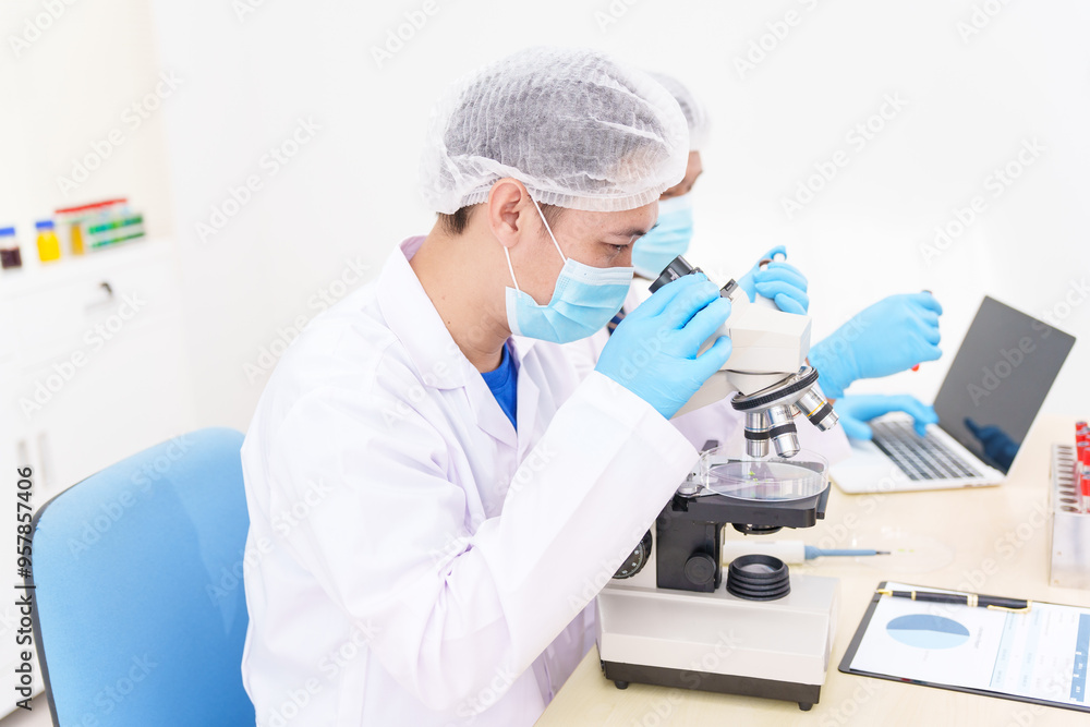 Two Asian scientists in lab coats sit at a table in a laboratory, examining samples through a microscope. The room is equipped with scientific tools, liquids, and chemistry materials. Two Asian scientists in lab coats sit at a table in a laboratory, examining samples through a microscope. The room is equipped with scientific tools, liquids, and chemistry materials.