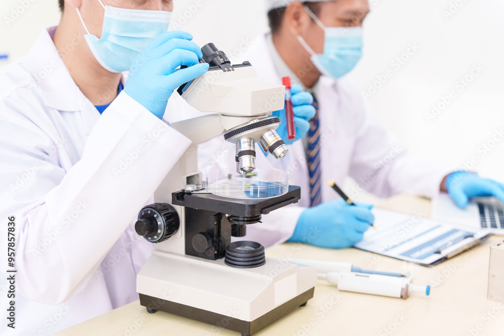 Two Asian scientists in lab coats sit at a table in a laboratory, examining samples through a microscope. The room is equipped with scientific tools, liquids, and chemistry materials. Two Asian scientists in lab coats sit at a table in a laboratory, examining samples through a microscope. The room is equipped with scientific tools, liquids, and chemistry materials.