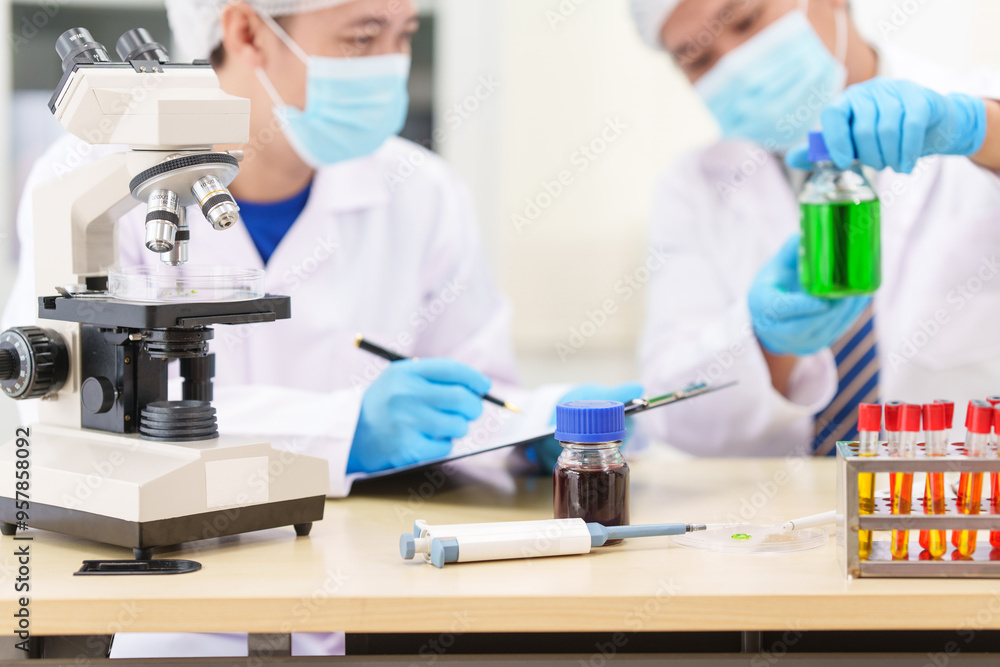 Two Asian scientists in lab coats sit at a table in a laboratory, examining samples through a microscope. The room is equipped with scientific tools, liquids, and chemistry materials. Two Asian scientists in lab coats sit at a table in a laboratory, examining samples through a microscope. The room is equipped with scientific tools, liquids, and chemistry materials.