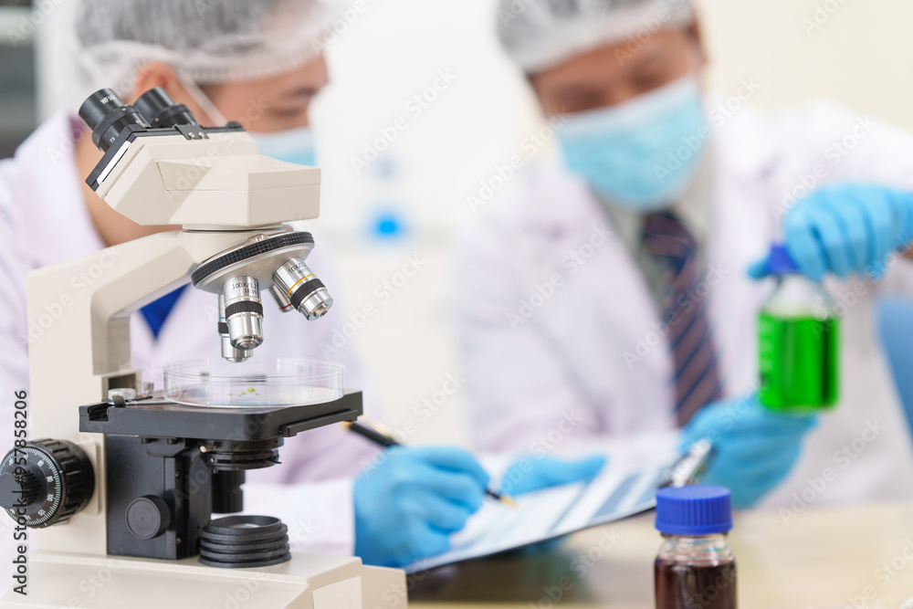 Two Asian scientists in lab coats sit at a table in a laboratory, examining samples through a microscope. The room is equipped with scientific tools, liquids, and chemistry materials. Two Asian scientists in lab coats sit at a table in a laboratory, examining samples through a microscope. The room is equipped with scientific tools, liquids, and chemistry materials.