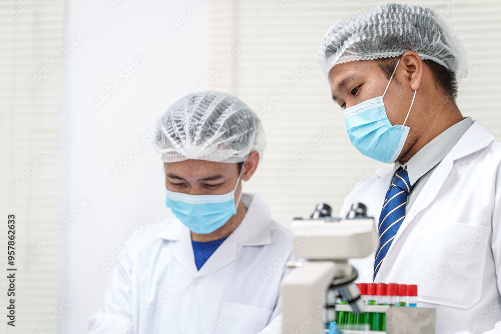 Two Asian scientists in lab coats sit at a table in a laboratory, examining samples through a microscope. The room is equipped with scientific tools, liquids, and chemistry materials. Two Asian scientists in lab coats sit at a table in a laboratory, examining samples through a microscope. The room is equipped with scientific tools, liquids, and chemistry materials.