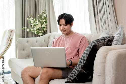 An Asian man in casual clothes using his laptop on a couch in the living room, working remotely from home or scrolling on website.