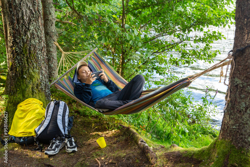 A teenage boy is relaxing in a hammock on the shore of a forest lake and playing with a smartphone.