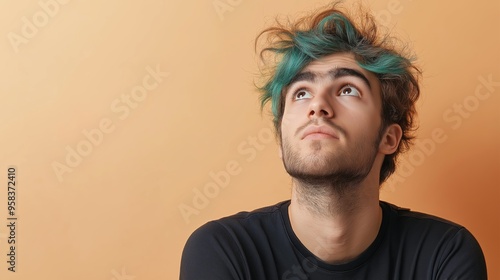 A mid-20s Caucasian man with teal quiff hair looks up curiously against a light peach background, wearing a black t-shirt