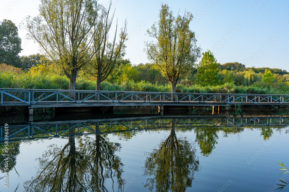 A bridge over a pond with trees in the background. The water is calm and the reflection of the trees and bridge can be seen in the water A bridge over a pond with trees in the background. The water is calm and the reflection of the trees and bridge can be seen in the water