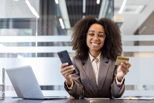 Portrait of a smiling young African American woman in a suit sitting in the office at a desk, holding a phone and a credit card, looking at the camera