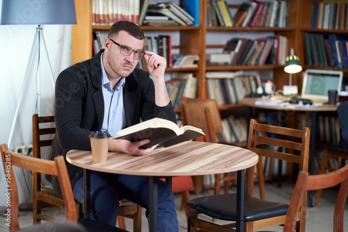 Man reading book and drinking coffee in a library cafe with many books on a background