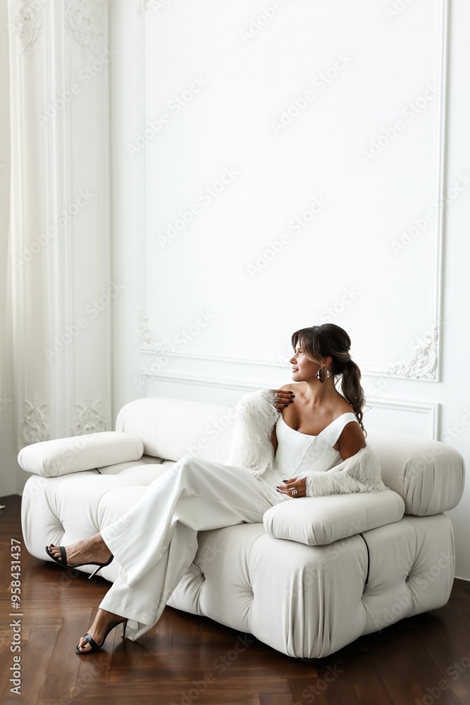 Beautiful woman model in white sitting on white sofa in light room. Chic brunette woman wearing a white suit leans casually against a white piece of furniture in a pristine studio. Beautiful woman model in white sitting on white sofa in light room. Chic brunette woman wearing a white suit leans casually against a white piece of furniture in a pristine studio.