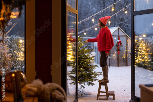 Young woman in red decorates lush Christmas tree with festive ballls and garland at backyard of her house on snow fall, preparing for a winter holidays photo