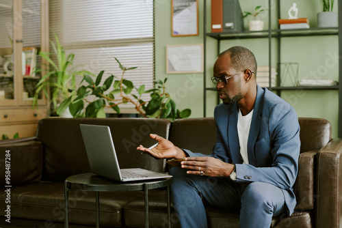 Bilde på lerret Man wearing suit and glasses conducting virtual meeting in a modern office setting while seated on brown sofa