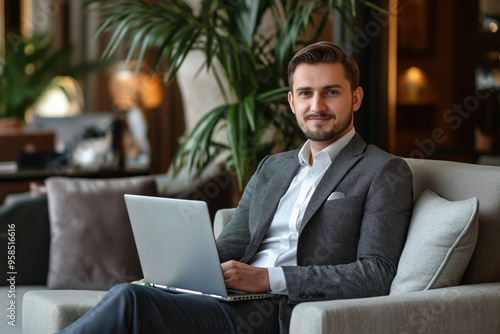 A man in a suit is sitting on a couch with a laptop in front of him. He is smiling and he is enjoying his time