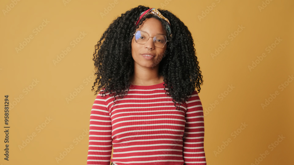 Young woman with curly hair in glasses wearing striped sweater looking at camera listening carefully to her interlocutor standing on yellow background in studio Young woman with curly hair in glasses wearing striped sweater looking at camera listening carefully to her interlocutor standing on yellow background in studio