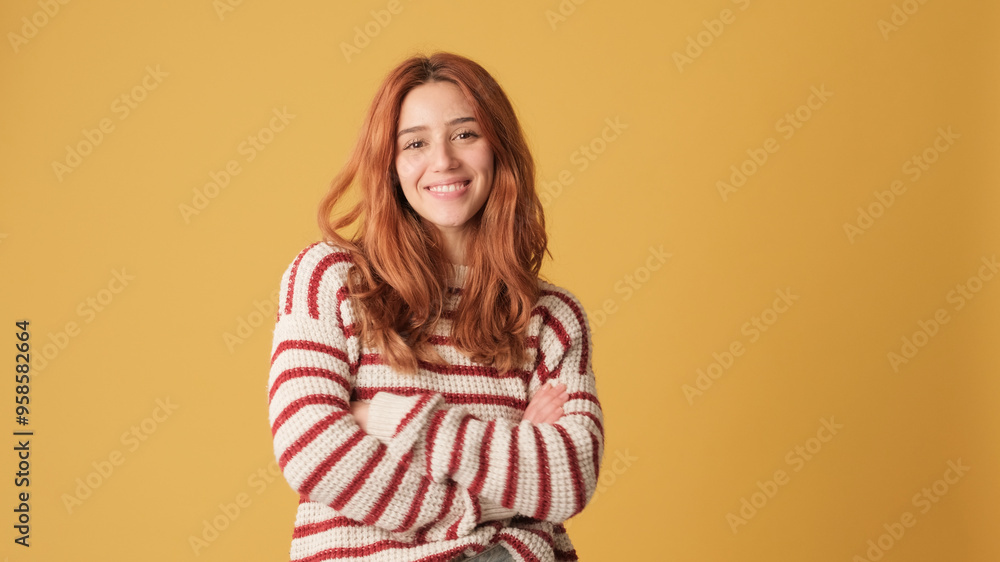 Smiling red-haired woman dressed in sweater crosses her arms looking at camera on yellow background. Beautiful girl poses in studio with blank space on yellow wall for your promotion Smiling red-haired woman dressed in sweater crosses her arms looking at camera on yellow background. Beautiful girl poses in studio with blank space on yellow wall for your promotion