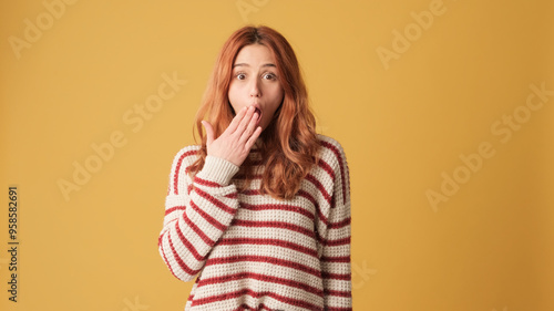 Surprised red-haired woman dressed in sweater looking at camera with big eyes, covering her mouth with her palm and looking to side on yellow background